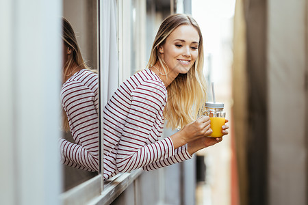 微笑的女人喝着一杯天然的橙汁 从她家窗外倾斜而下黑发高清图片素材