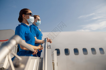 护士礼仪素材两名穿蓝制服的年轻空姐 太阳镜和防护面罩的侧视 在登机时站在飞机上站立着背景