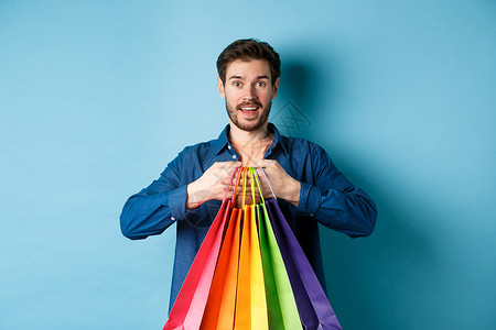 快乐的年轻人拿着色彩多彩的购物袋 微笑兴奋 买礼物 站在蓝背景上工作室情绪男人购物成人广告购物者店铺促销商业背景图片