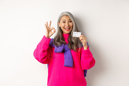 购物概念 微笑的亚洲中年女性 头发灰白 展示塑料信用卡和 OK 标志 推荐银行促销 白色背景祖母成人喜悦广告化妆品奶奶乐趣卡片母背景图片