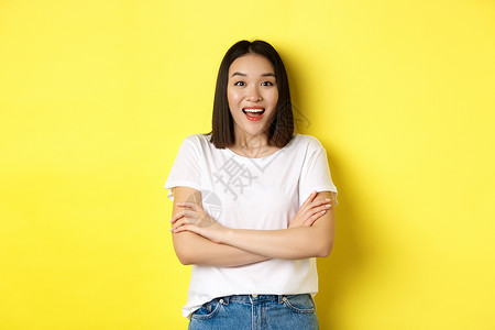 美丽和时尚的概念 快乐的亚洲女性穿着白色T恤衫 胸前交叉手臂 对着镜头大吃一惊 站在黄色背景上女孩黑发促销工作室理发发型学生广告背景