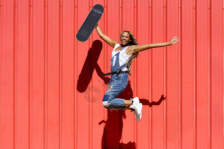 黑女人穿着随随便便的衣着 Wtih 一个滑板跳跃 快乐地跳在红色的城市墙壁背景街道编织幸福青年女孩黑发女性发型女士恐惧背景图片