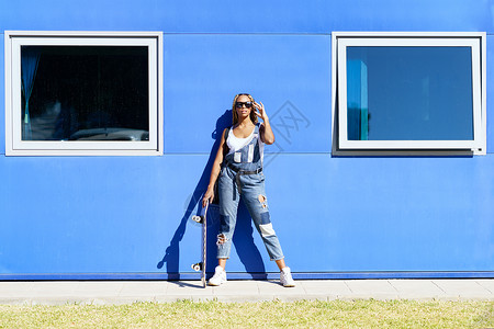 黑女人穿着随随便便的衣服 穿着现代太阳镜和蓝墙底的滑板背景图片