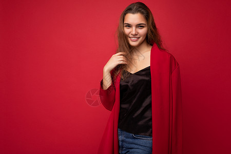年轻漂亮的欧洲时尚黑发女人身穿黑色上衣和红色开衫 在红色背景中显得格格不入 带着积极真诚的情感 看着相机简单自然 可用空间衬衫女背景图片