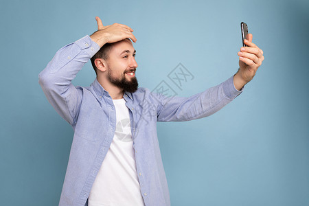 身着临时时装服装的英俊年轻黑发胡子男子 在蓝色背景墙上与世隔绝 手持智能手机 用自拍照片观看移动电话屏幕显示t恤神器博客电话摄影背景图片