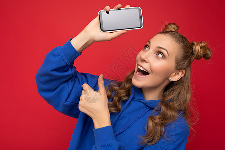 身穿红色背景中突显的时尚蓝色连帽衫 面带微笑 面带微笑的迷人金发女郎 手握智能手机的复印空间显示手机 空屏显示指向 gadjet背景图片