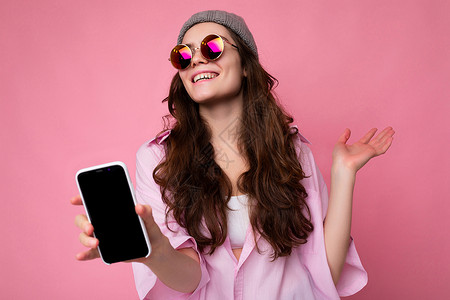 正面微笑 迷人的黑发年轻女性 戴着时尚的粉色衬衫灰色帽子 戴着彩色太阳镜 在粉色背景中显得格格不入 手里拿着手机 展示着空显示屏背景图片