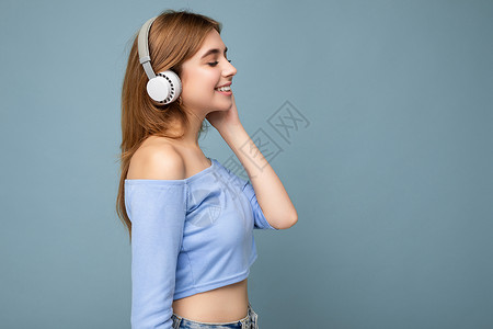 蓝色耳机素材侧面照片是美丽正面微笑的年轻金发女子身穿蓝色裁剪上衣 与蓝色背景墙隔开 头戴白色无线蓝牙耳机 听着酷炫的音乐 看着旁边背景