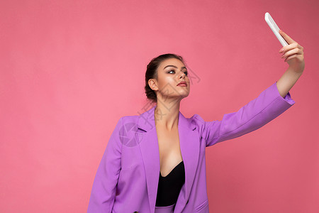 照片中 身穿紫色西装的漂亮年轻女子在手机上自拍 粉红色背景中 她正看着手机摄像头星星社交神器社会电话摄影套装知名度屏幕相机背景图片
