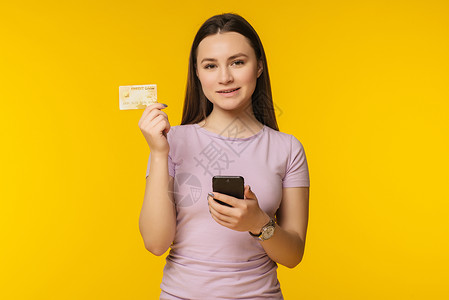 一个快乐的金发年轻女孩的肖像 她展示塑料信用卡 却持有黄色背景隔绝的手机-图像背景图片