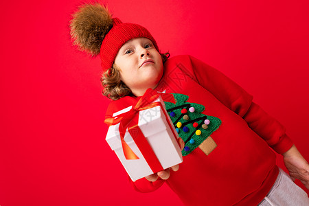 一个红墙背景卷发的酷男孩 穿着毛衣 手里拿着圣诞树 手里拿着礼盒魅力购物情绪销售新年礼物盒子红色背景图片
