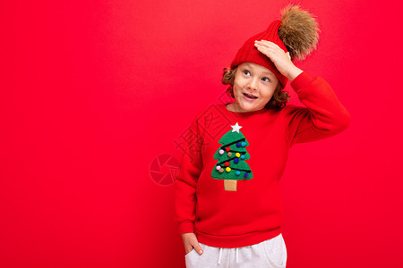 喜笑的男孩在红色背景 在温暖的帽子和毛衣 与圣诞树销售情绪卷发新年行动背景图片