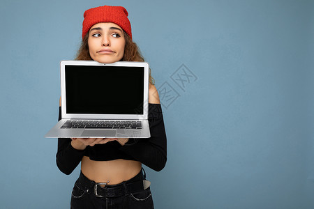 浅蓝色上衣美丽有趣的年轻深色卷发女孩穿着黑色裁剪上衣和红色和橙色的 do-rag 隔离在浅蓝色墙壁背景上 拿着电脑笔记本电脑 屏幕显示器空背景
