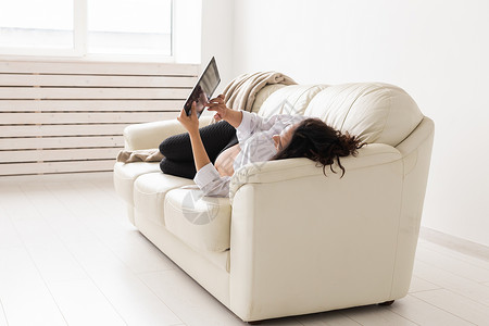 西班牙裔怀孕妇女在家里躺在沙发上时使用平板电脑 怀孕和为人父母的概念信息 母亲的姓氏阅读女孩腹部肚子享受电子书女性拉丁技术产妇背景图片