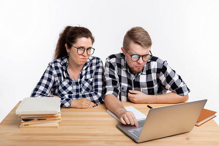 人与教育概念两名穿着格格衬衫坐在桌边的令人迷惑的学生逗乐笔记本夫妻图书工作室眼镜电脑极客成人享受背景图片