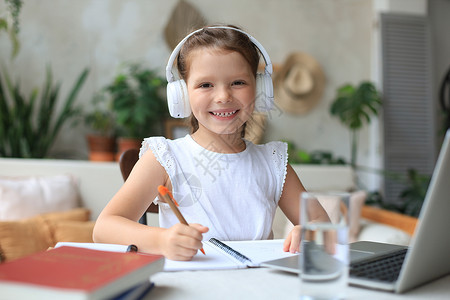 电脑pC戴着耳机的微笑小女孩在家里使用笔记本电脑在线学习手写 戴着耳机的可爱快乐的小孩在 PC 上上互联网课程或课程书本工作生活方式屏幕背景