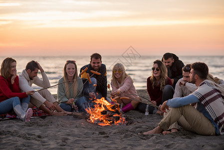 坐在海滩火炉边的年轻朋友团体饮料营火烧烤细胞火焰假期友谊旅游野餐乐趣背景图片