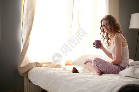 年轻的美女坐在床上 用笔记本电脑黑发咖啡面包卧室日记微笑枕头杯子羊角字帖背景图片