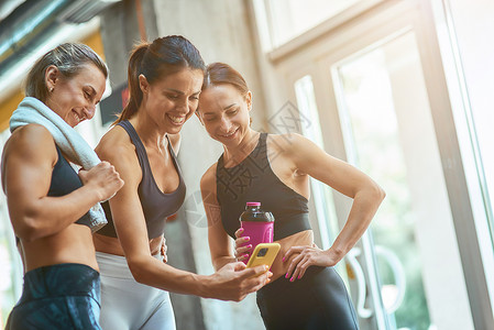 三名年轻美丽快乐的运动女性在健身房锻炼后休息时在智能手机上自拍 一起锻炼背景图片