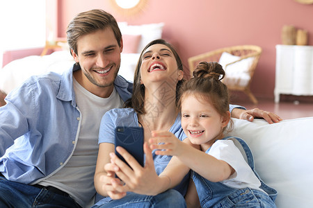 家庭电话素材美丽的年轻家庭 孩子在沙发上拿着自拍 家里有智能手机的女孩童年女士技术乐趣照片微笑电话时间母亲帽子背景