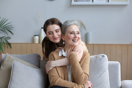 快乐的女儿拥抱年老的母亲 坐在沙发后面的客厅里 在家享受温柔的时光妈妈父母老年黑发女士成人笑脸家庭祖母咖啡背景