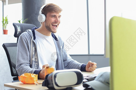使用电脑 流线播放或漫步视频 微笑着穿便衣的年轻男子玩家耳机编码技术专注黑客男性虚拟现实小吃男人背景图片