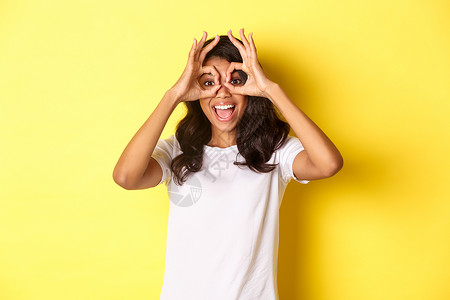 美籍非洲女孩的快乐和有趣的形象 制作手指眼镜 透过它们看一些令人惊叹的东西 站在黄色背景上站立发型广告手势员工多样性成人工作室女背景图片