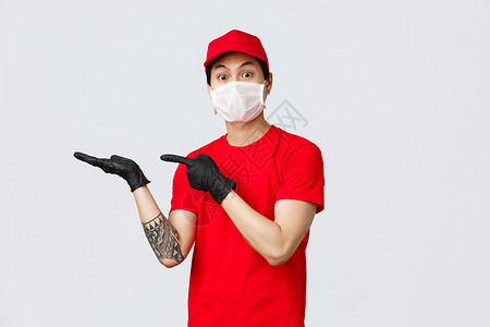 看这个 身穿红帽 T 恤 戴着防护乳胶手套和医用口罩的兴奋的亚洲送货员 手里拿着一些东西 指着手掌 盯着镜头印象深刻杂货店餐厅房背景图片