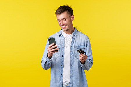 技术 生活方式和广告概念 帅气的微笑快乐的金发男人看着手机应用程序 在购物网站上支付订单或送货 持有信用卡 黄色背景背景图片