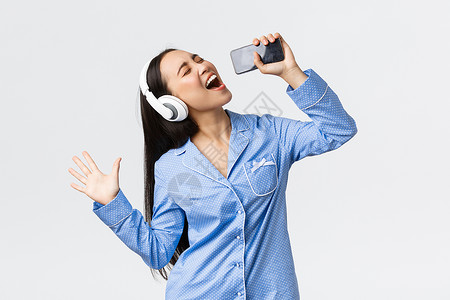 音乐列表居家休闲 周末和生活方式的概念 穿着睡衣的兴奋而无忧无虑的亚洲女孩 在智能手机上播放卡拉 OK 应用程序 戴着耳机在手机里唱歌背景