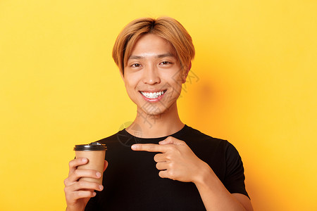 最接近满意的英俊金发亚裔金发男子 笑笑喜悦和手指指着美味的咖啡杯 站在黄色背景上情感男朋友促销成人广告职业学生员工教育理发背景图片