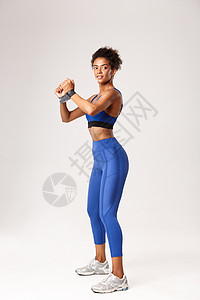 运动蓝色背景身穿蓝运动服的有吸引力的非洲-美国健身女孩全长照片 用橡皮带做锻炼和看远视 在白种背景下打工背景