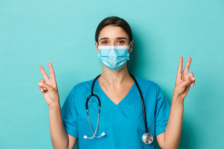 医用标志covid-19 的概念和检疫概念 戴着医用面具和磨砂膏的开朗迷人的女医生 展示和平标志 站在蓝色背景上背景