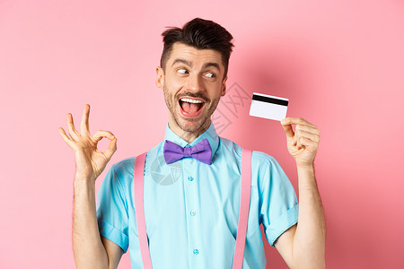 欢欣鼓舞的人建议伟大的宣传 显示OK标志和看着兴奋的塑料信用卡 站在粉红背景上成人工作室男人银行吊带裤胡子促销购物领结快乐背景图片
