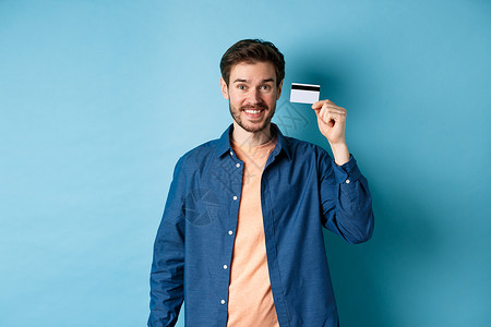 快乐的caucacians人笑着微笑 展示塑料信用卡 穿着蓝色背景的便衣站立胡子购物者金融发型成功成人办公室生活男人工作室背景图片