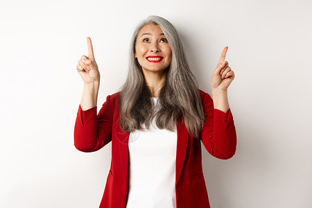 穿着红色西装外套和化妆的快乐亚洲女士 看着并指着手指 展示特别优惠 站在白色背景上办公室商务退休技术头发广告工人商业成人人士背景图片