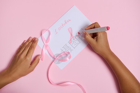 十月1号一位女士手握毡尖笔的顶部视图写下了 10 月 1 日 并在纸上画了一个粉红色的乳腺癌宣传月标志 一条粉红色的丝带 末端无尽 躺在背景