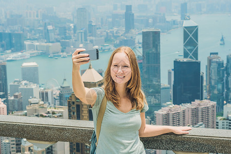 凌霄塔香港太平山女人用智能手机拍自拍杆照片 欣赏维多利亚港的景色 香港凌霄阁顶部的观景台 散焦背景 旅游亚洲概念女士港口观光天际城市景背景