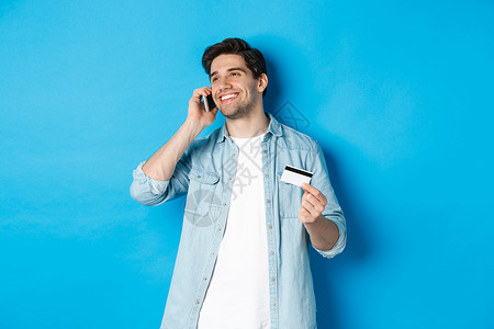 微笑的男人打电话给银行支持 在手机上聊天 拿着信用卡 站在蓝色背景下商业广告胡子黑发订金工具促销购物细胞网络背景图片