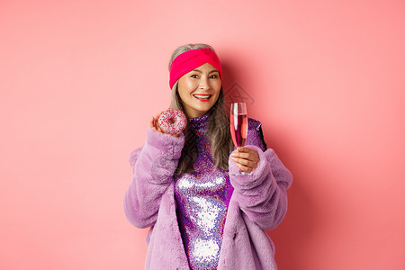 甜甜圈促销主图穿着紫花皮大衣 吃甜甜甜圈 喝香槟 在派对上玩得开心 站在粉红色背景之上的优美和时尚的女性促销乐趣成人头巾化妆品食物购物快乐广告背景