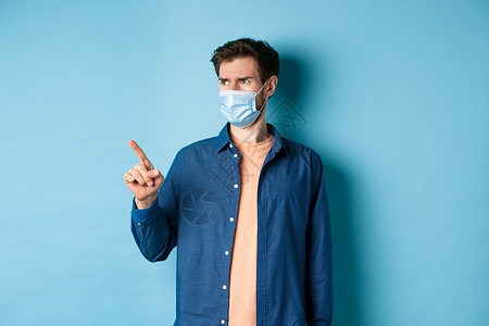 Covid19 和医疗保健概念 戴着面罩的困惑而怀疑的男人皱着眉头 指着 看着空荡荡的空间 站在蓝色背景上情绪面具成功社交广告促背景图片