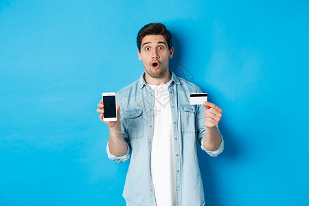 惊讶的年轻人展示手机屏幕和信用卡 在网上购物 站在蓝色背景下成人胡子促销潮人广告技术商业微笑贷款银行背景图片