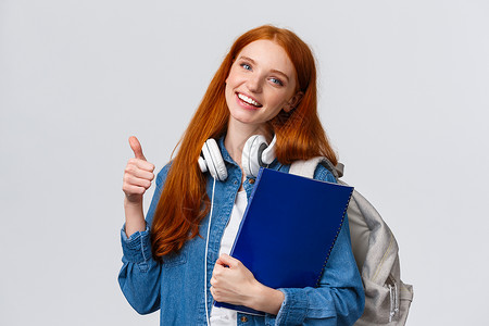 教育 青少年和学生生活方式的概念 快乐的红发可爱女孩背着背包和文件夹 把她的作品集带到艺术 设计课上 竖起大拇指表示赞同 比如手背景图片
