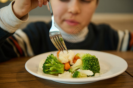 拿着叉子男孩关注一盘蒸菜 健康的花椰菜和胡萝卜餐 防止模糊的男孩坐在桌边吃东西并举着叉子 营养 为幼儿做素食背景