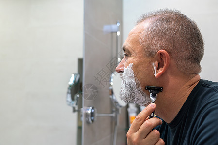长着英俊白发的白头发熊男子在照镜子时刮胡子 改变风格的概念头发胡子男性剃刀凝胶中年奶油胡须泡沫隔断背景图片