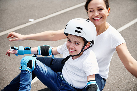 戴着防护头盔和装备的快乐妈妈和可爱的儿子坐在滑板上享受时光背景图片
