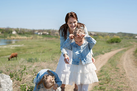 母亲和她的双胞胎女儿走在靠近奶牛的村庄的一条土路上 母女俩扮牛玩得不亦乐乎小屋游戏草地家庭森林旅行快乐喜悦呵护姐姐背景