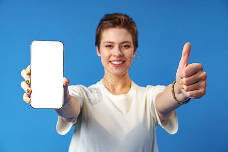 一名年轻女性在蓝背景上站立时展示空白屏幕移动电话的纵向肖像手机相机女士手指微笑女孩细胞广告成人营销背景图片
