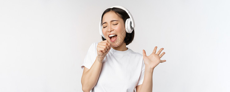 白色耳机韩国女孩在耳机中唱歌和听音乐 玩得开心 站在白色背景之上互联网工作室横幅购物企业家女性学生女士成人女朋友背景