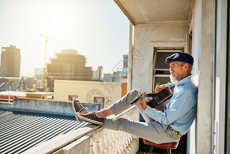 他弹吉他的日常仪式 拍摄一位和平的老人坐在他的阳台上 同时在背景中弹奏原声吉他与城市背景图片
